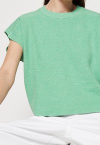 Kvinna iklädd en ljusgrön kortärmad stickad topp tillsammans med vita byxor mot en enfärgad bakgrund.