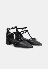 Tacones de cuero negro con punta afilada y un pequeño tacón cuadrado. Presenta múltiples correas con detalles de herrajes dorados. Textura suave.
