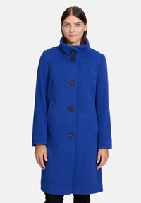 Kobieta ubrana w krótką, królewską niebieską płaszcz do kolan z wysokim kołnierzem i czterema czarnymi guzikami, stojąca na tle białym.