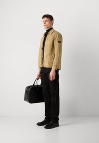 Veste légère beige avec une finition texturée, col roulé noir, pantalon noir et sac en cuir noir à la main ; se tenant devant un fond neutre.