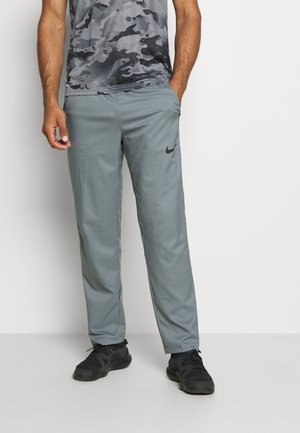 Pantalons de sport gris en tissu lisse et léger, avec des poches latérales et un petit logo noir sur la cuisse gauche. Portés avec des baskets noires.