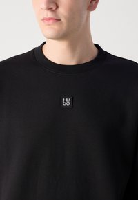 Sudadera negra con cuello redondo, que cuenta con un parche de logo texturizado etiquetado "HUGO" en el pecho. Tejido suave con un corte relajado.
