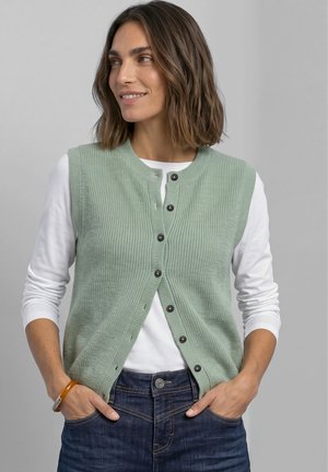 Femme aux cheveux bruns mi-longs portant un gilet en tricot vert sauge boutonné sur une chemise blanche à manches longues et un jean bleu, les mains dans les poches.