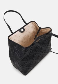 Sac cabas en cuir matelassé noir avec motifs de logo embossés, doublure intérieure beige imprimée avec "GUESS," et fermeture magnétique.