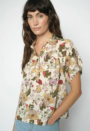 Žena s dlhými hnedými vlasmi, oblečená v krémovej blúzke s krátkym rukávom a farebným kvetinovým vzorom, a svetlomodrých džínsoch, s rukou v kapse.