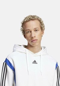 Haut à capuche blanc avec des accents rayés bleus et noirs. Le matériau semble doux ; présente le logo Adidas sur la poitrine et des cordons de serrage sur la capuche.