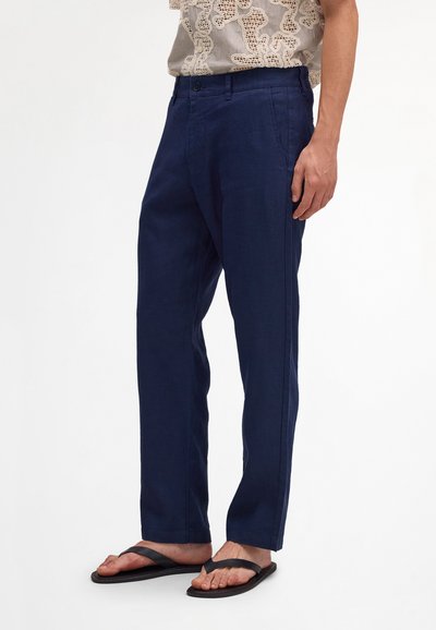 Mężczyzna ubrany w granatowe spodnie, beżową koszulę w wzory i czarne klapki, stojący na białym tle.