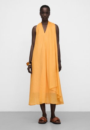 Vrouw draagt een mouwloze oranje midi-jurk met asymmetrische drapering, bruine slippers en chunky armbanden van schildpadmotief aan haar linkerpols.