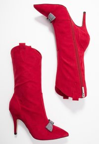 Bottes à talons hauts en suède rouge avec des bouts pointus et un accent de nœud décoratif. Dotées d'une fermeture éclair sur le côté et d'un design ajusté et moulant.