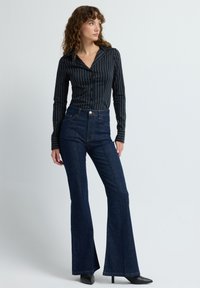 Černobíle pruhovaná košile na knoflíky spárovaná s tmavými džínami s vysokým pasem a rozšířenými nohavicemi, s kontrastním šitím a jemným leskem.