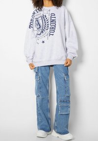 Sudadera gris con un estampado gráfico que presenta "Toronto" y un diseño de insignia. Combinada con jeans cargo azules y zapatillas blancas.