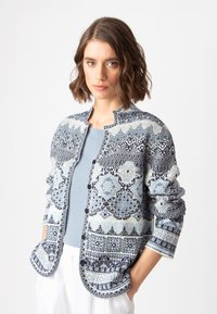 Zils un balts adīts kardigāns ar sarežģītiem ģeometriskiem rakstiem, apaļš dekoltē, pogas un kabatas, uzvilkts virs zila krekla.