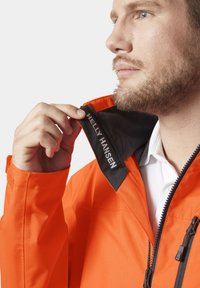 Pomarańczowa wodoodporna kurtka Helly Hansen z czarnym kołnierzem. Posiada zamek z przodu, teksturę materiału oraz logo wewnątrz kołnierza.