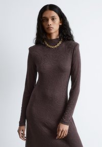 Braunes Langarmkleid mit Paisleymuster, hohem Kragen und strukturiertem Stoff. Auffällige Kettenhalskette sorgt für einen Kontrast.