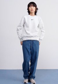 Grijze sweatshirt met ronde hals en zwart Nike-logo, gecombineerd met vervaagde blauwe jeans en lichtgrijze sneakers, staande op een blauwe vloer.