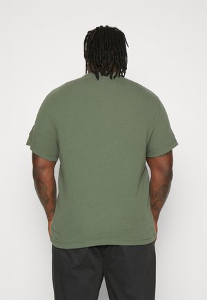 Olivgrünes T-Shirt mit kurzen Ärmeln, einer strukturierten Oberfläche, rundem Ausschnitt und aufgekrempelten Ärmeln. Kein sichtbares Branding oder grafische Details.