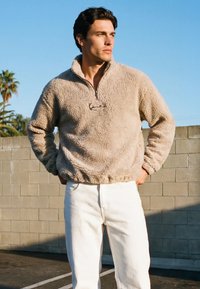 Egy férfi áll a szabadban, bézs polár pulóvert és fehér nadrágot visel, kezét zsebre dugva, felette napsütéses égbolt.