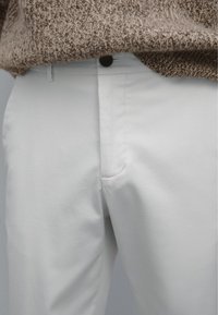 Witte katoenen broek met een platte voorkant, voorzien van een zichtbare zak en een zwarte knoopsluiting, gecombineerd met een getextureerde bruine trui.