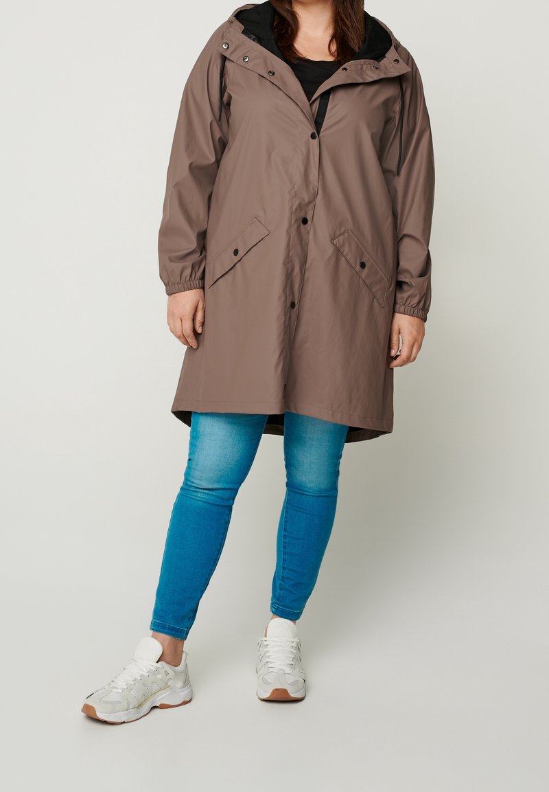 Personne portant un imperméable marron à capuche, un jean bleu et des baskets blanches, debout devant un fond uni clair.