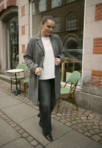 Grau karierten Mantel, weißes Oberteil, graue Jeans und schwarze Knöchelstiefel. Außensetting mit grün gemusterten Stühlen und Kopfsteinpflaster.