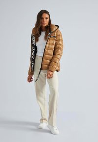 Giubbotto puffo marrone con finitura lucida, caratterizzato da una zip nera e un audace logo bianco dettagliato sulla parte anteriore, abbinato a pantaloni beige chiaro.