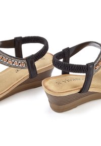 Schwarze Keilsandaletten mit einem elastischen Riemen und dekorativen Akzenten. Weicher beigefarbener Fußbett mit einer strukturierten braunen Außensohle.