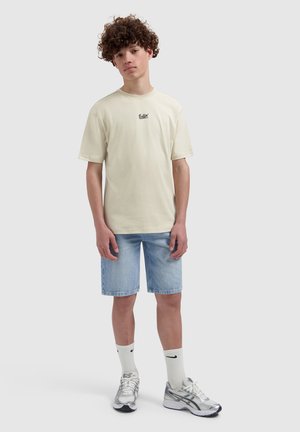 Teenager with curly hair wearing a beige "Ballin'" T-shirt, knee-length denim shorts, white Nike socks, and silver running shoes.
