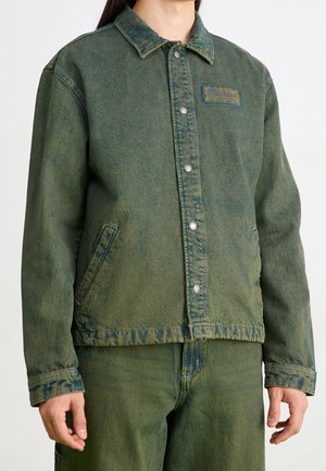 Personne portant une veste en denim verte avec des boutons-pression et un pantalon en denim vert assorti, debout devant un fond uni.