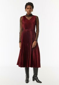 Burgunderfarbenes plissiertes Kleid mit tiefem V-Ausschnitt und transparenten schwarzen langen Ärmeln, kombiniert mit knielangen schwarzen Blockabsatzstiefeln.