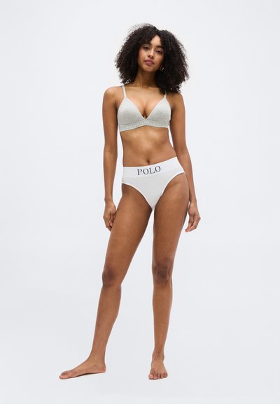Γυναίκα με κυματιστά μαλλιά που φοράει ανοιχτογκρι μπραλέτα και λευκά εσώρουχα με ψηλή γραμμή με ζώνη μέσης "POLO", όρθια ξυπόλυτη σε λευκό φόντο.