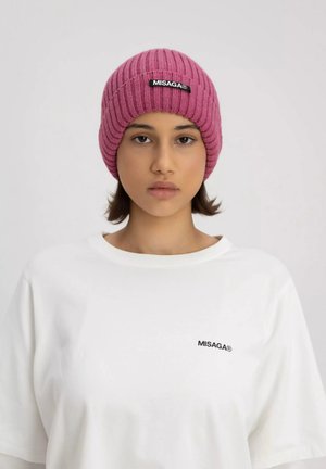 Roze geribbeld gebreid beanie met een zwart logo patch, gedragen met een witte t-shirt met korte mouwen met een klein zwart logo op de borst.