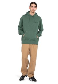 Joven de pie con las manos en los bolsillos, vistiendo un sudadera verde, pantalones beige y zapatillas negras frente a un fondo blanco.