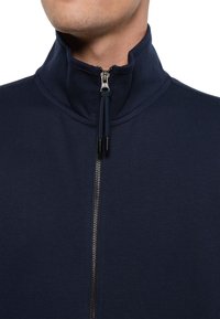 Marineblauer Reißverschlussjacke mit hohem Kragen, ausgestattet mit einem Metallreißverschluss auf der Vorderseite und strukturierter Stoffoberfläche. Akzente bilden die schwarzen Kordelzugenden.