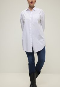 Camisa blanca de botón con bolsillo en el pecho, mangas largas y dobladillo redondeado, combinada con jeans azul oscuro y botas negras de estilo vaquero.