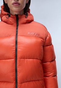 Světlá oranžová puffová bunda s kapucí, která má zipové uzávěr a rozšířené prošívané části. Materiál má lesklou texturu s logem na hrudi.