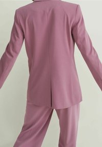 Vue de dos d'une personne portant une veste de costume ample mauve et un pantalon large assorti, sur un fond clair uni.