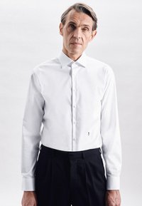 Witte langemouwen overhemd met een puntkraag, met een klein bloemembleem op de linkerzoom, gecombineerd met donkere geplooide broeken.