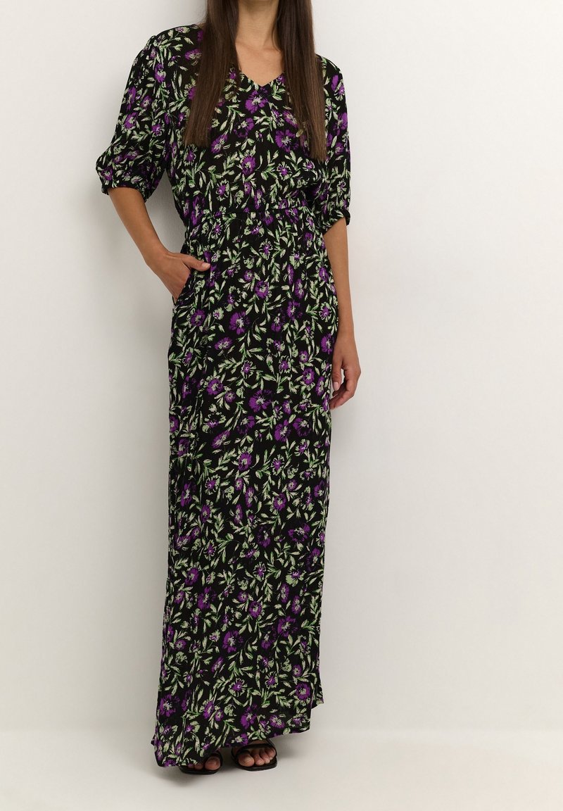 Femme portant une robe longue noire à imprimé floral violet et vert, main dans la poche, debout devant un mur clair uni.
