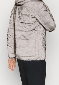 Mand i let grå digital camo-mønstret puffet jakke og sorte bukser, vist fra ryggen og siden mod en ensfarvet baggrund.
