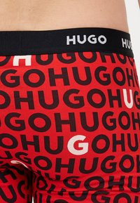 Boxers rouges avec un motif répété en noir et blanc du logo "HUGO", dotés d'une ceinture élastique noire affichant "HUGO" en texte blanc.
