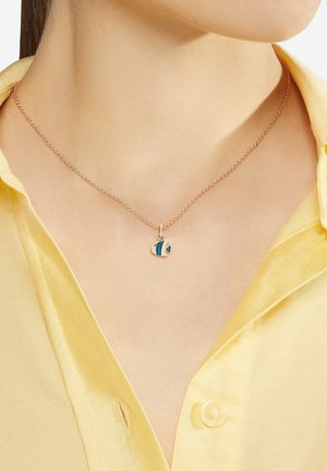 Zarte goldene Halskette mit kleinem, fischförmigem Anhänger in Blau und Gold an einer Person mit gelbem Hemd.