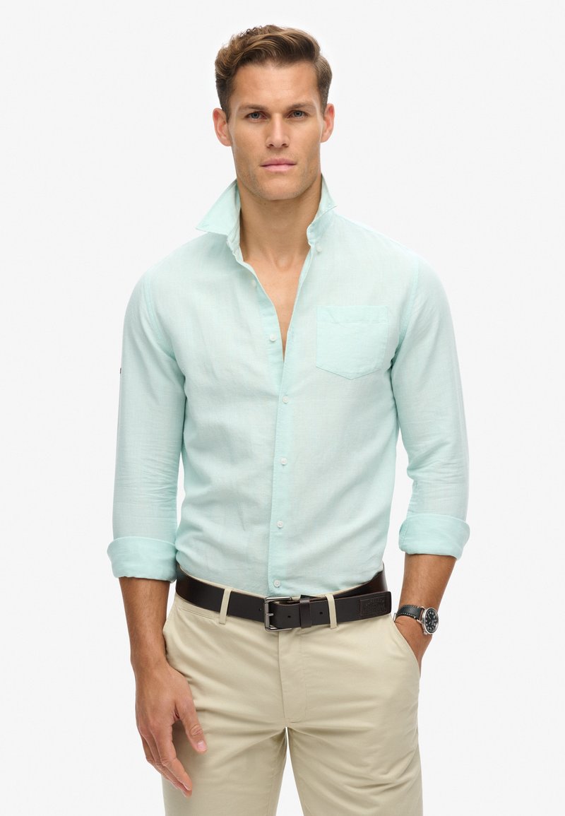Man wearing light blue button-up shirt with rolled sleeves, beige pants, black belt, and a wristwatch standing against white background.