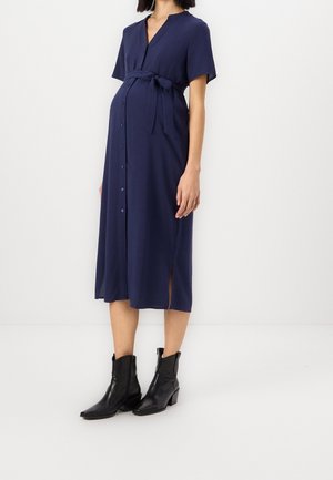 Femme enceinte portant une robe midi bleu marine boutonnée avec des manches courtes et des bottines noires, debout sur un fond blanc.