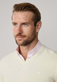 V-Ausschnitt Pullover aus creamfarbenem Stoff mit strukturierter Oberfl�che, kombiniert mit einem hellrosa Hemd mit Kragen darunter. Dezentes Logo auf der Brust.