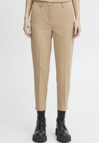 Beige broek met een op maat gemaakte pasvorm, voorzien van een gladde textuur, riemlussen, zijzakken en een taps toelopende broekspijp. Gecombineerd met zwarte laarzen.