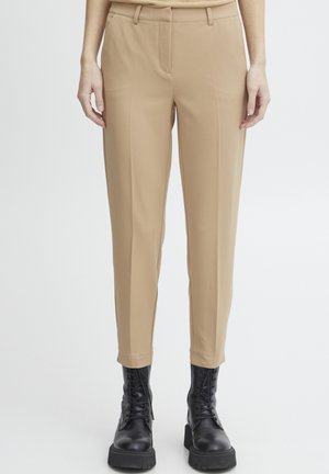 Pantaloni beige con vestibilità su misura, caratterizzati da una superficie liscia, passanti per cintura, tasche frontali e un design con gamba affusolata. Abbinati a stivali neri.