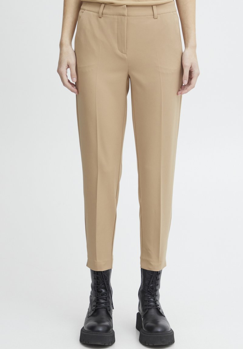 Beige broek met een op maat gemaakte pasvorm, voorzien van een gladde textuur, riemlussen, zijzakken en een taps toelopende broekspijp. Gecombineerd met zwarte laarzen.