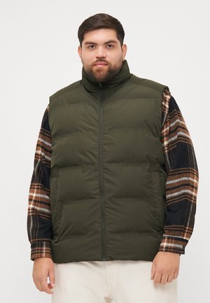 Olive green puffer vest with high collar, front zipper, and side pockets, worn over a multicolored plaid shirt.