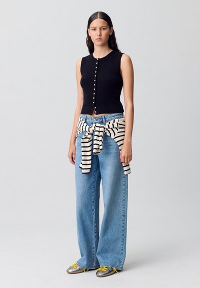 Top senza maniche nero con bottoni dorati, jeans a gamba larga blu chiaro e un maglione a righe bianco e nero legato in vita. Scarpe argento con lacci gialli.