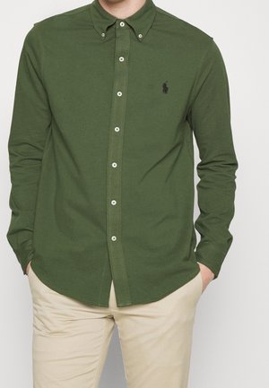 Mężczyzna ubrany w oliwkowozieloną koszulę na guziki z długim rękawem oraz beżowe spodnie, z rękami w kieszeniach, stojący na jednolitym tle.